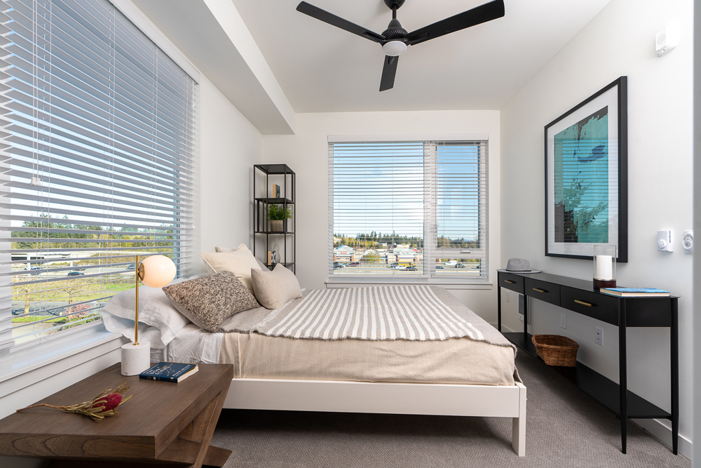 Minimalist bedroom with a large bed, beige bedding, bedside table with a lamp, black shelving unit, dresser, artwork on the wall, and large windows letting in natural light.