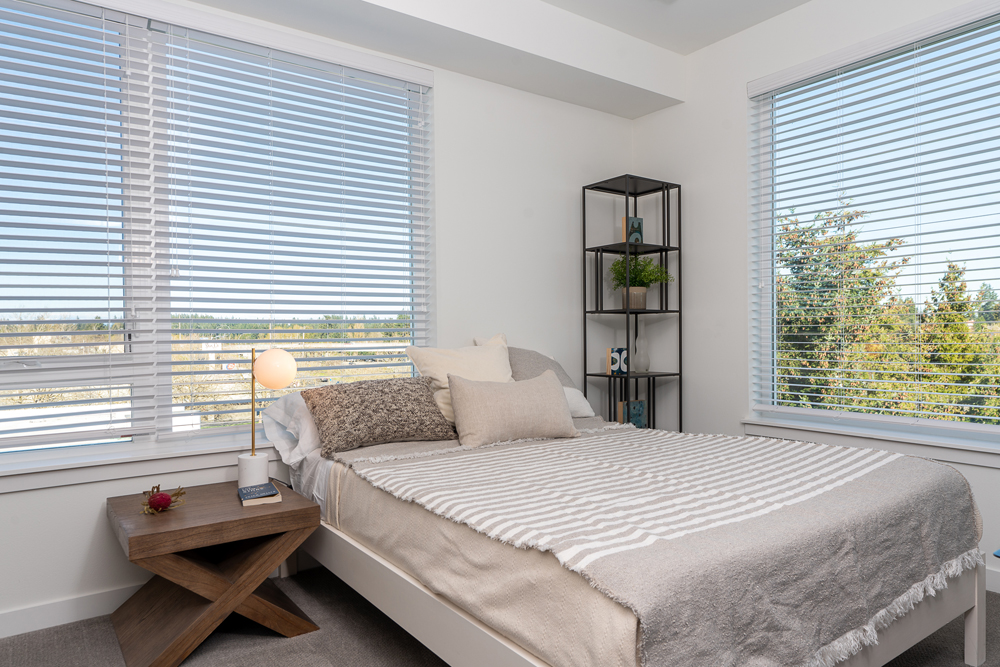 A bright bedroom with large windows, a neatly made bed with neutral bedding, a wooden nightstand with a lamp and small decor, and a metal shelf with plants and books in the corner. Trees are visible outside.