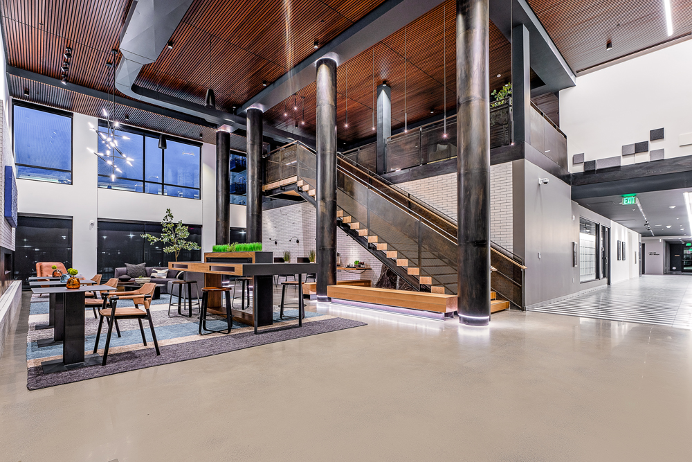 Modern lobby with tall black columns, wooden ceiling, a central staircase with glass railing, contemporary furniture, and large windows. Hallway and seating area visible, with plants and stylish lighting accents throughout.