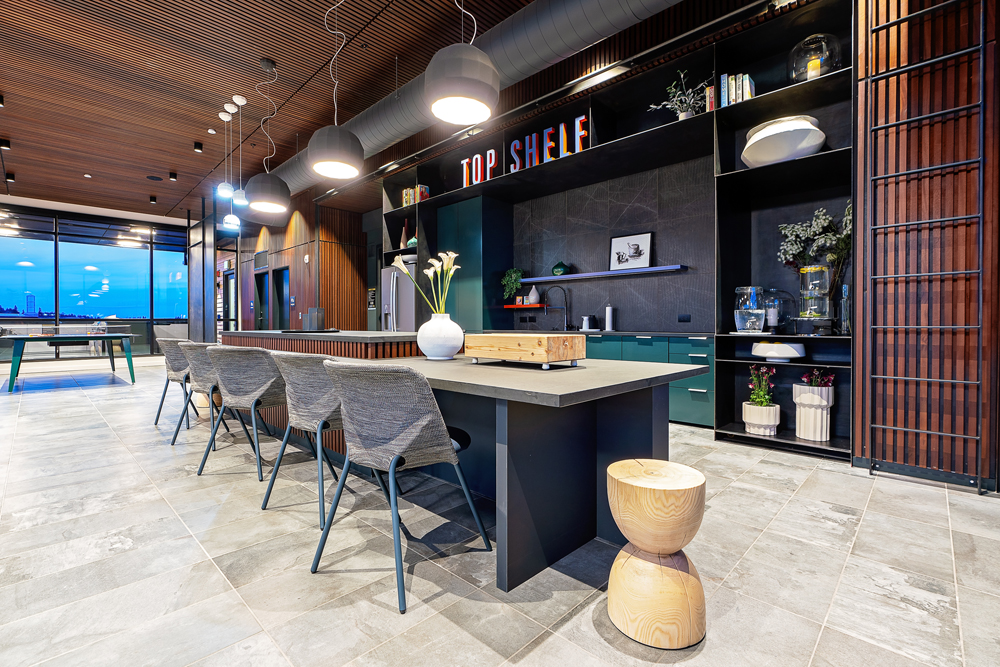 Modern communal kitchen and dining area with a long island table, four gray chairs, a wooden stool, pendant lighting, built-in shelves, decorative plants, and a sign reading TOP SHELF on the upper shelf.