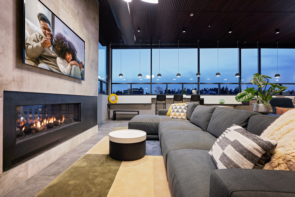 A modern living room with a large gray sectional sofa, pillows, a round coffee table, a fireplace, a green plant, and floor-to-ceiling windows showcasing a cityscape at dusk. A TV above the fireplace displays two people smiling.