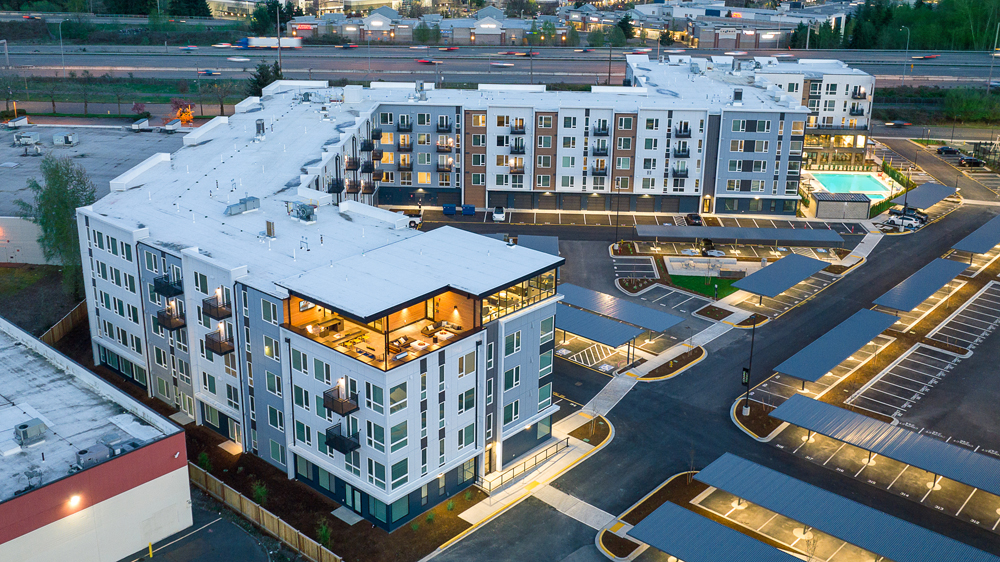 Aerial view of a modern, multi-story apartment complex with balconies, illuminated windows, parking lots, and a swimming pool, set near a busy highway and surrounded by other buildings.