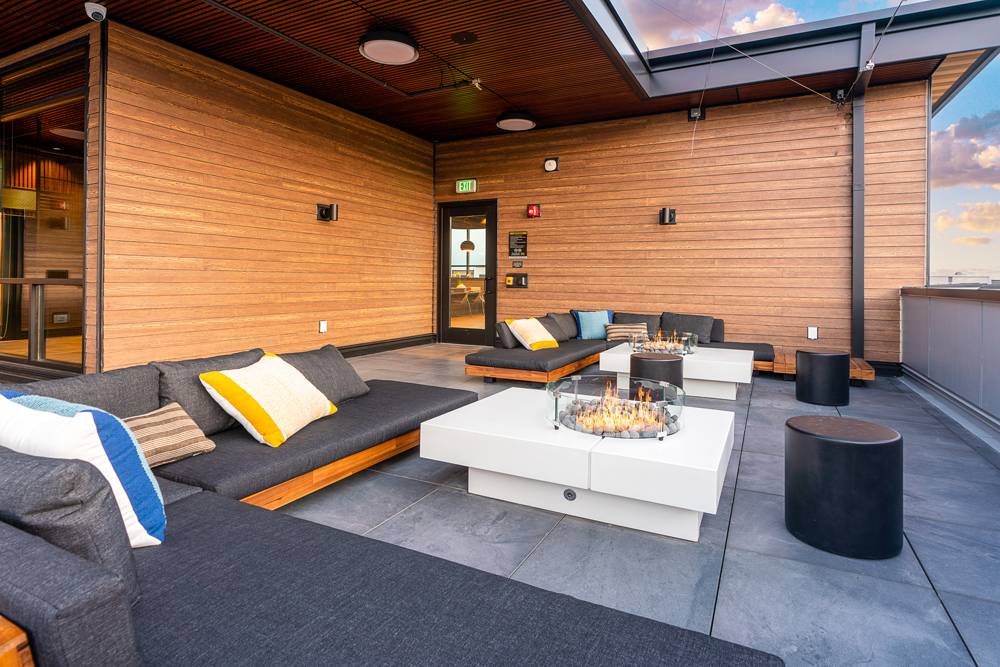Modern rooftop patio with wooden walls, cushioned seating, colorful pillows, and two square fire pit tables. The area has a relaxed, inviting atmosphere under a partially covered sky.