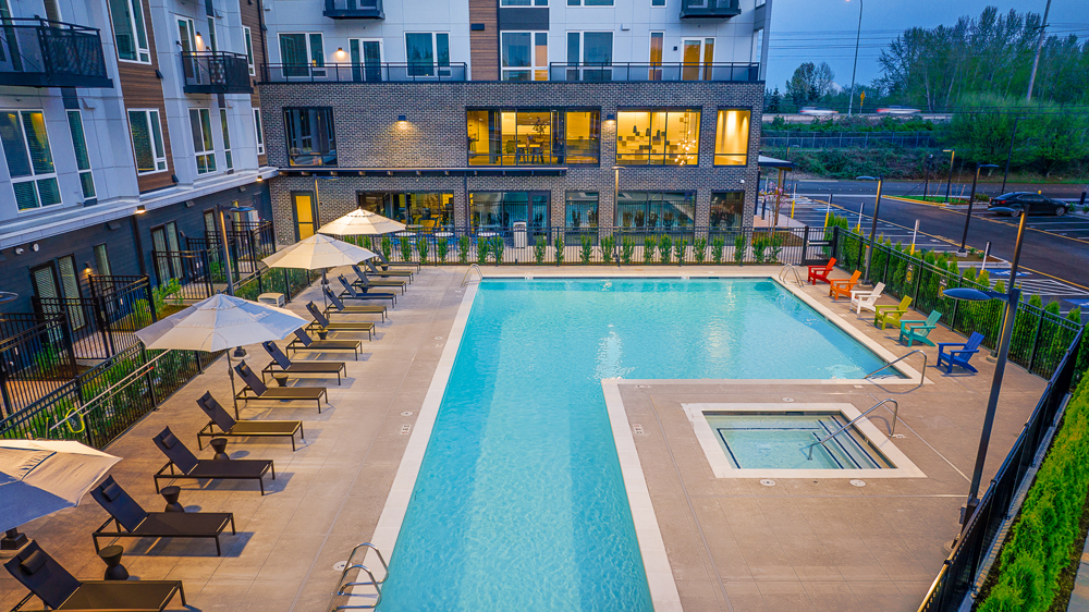 A modern apartment complex features an outdoor swimming pool with lounge chairs, umbrellas, and a hot tub. The area is surrounded by fencing, greenery, and the apartment building. Evening lighting creates a relaxed atmosphere.